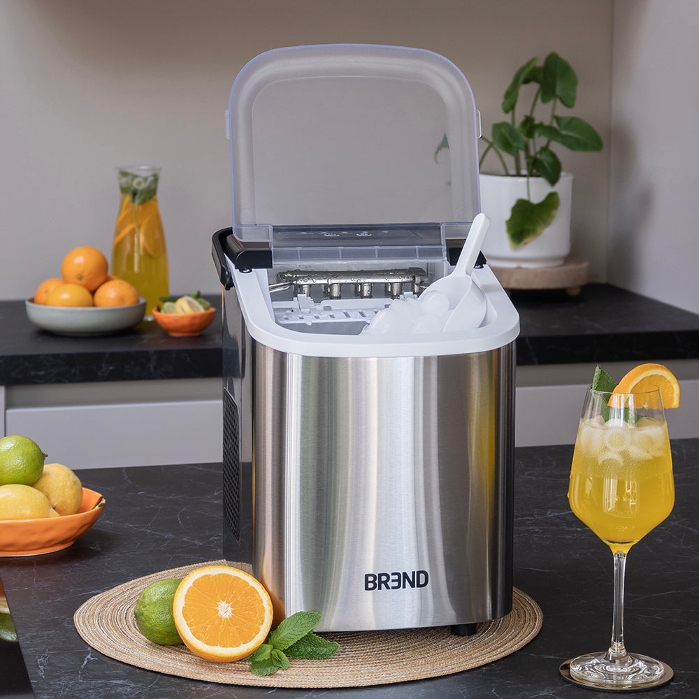 Schep vol ijsblokjes in de geopende roestvrijstalen ijsblokjesmachine naast een glas sinaasappeldrank