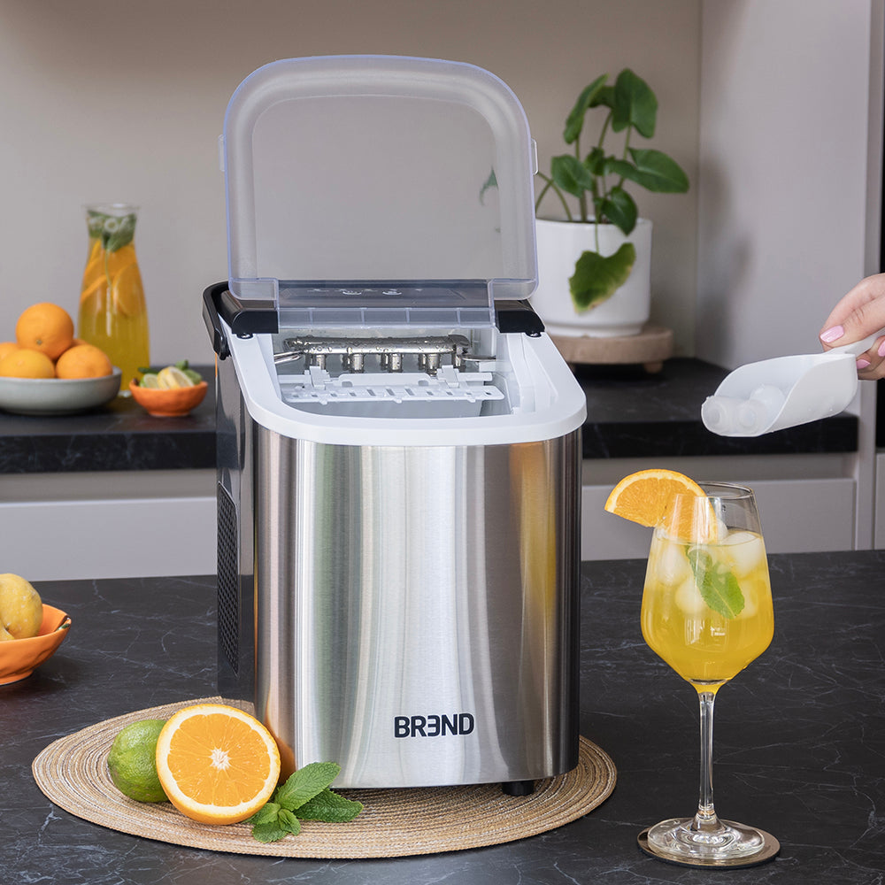 Hand schept ijsblokjes uit de roestvrijstalen ijsblokjesmachine naast een glas met sinaasappel