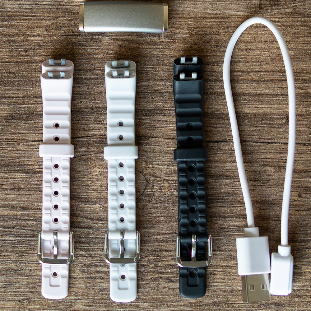 Activity tracker met drie verwisselbare bandjes in roze, lichtgrijs en zwart en een USB-oplaadkabel op een houten tafel