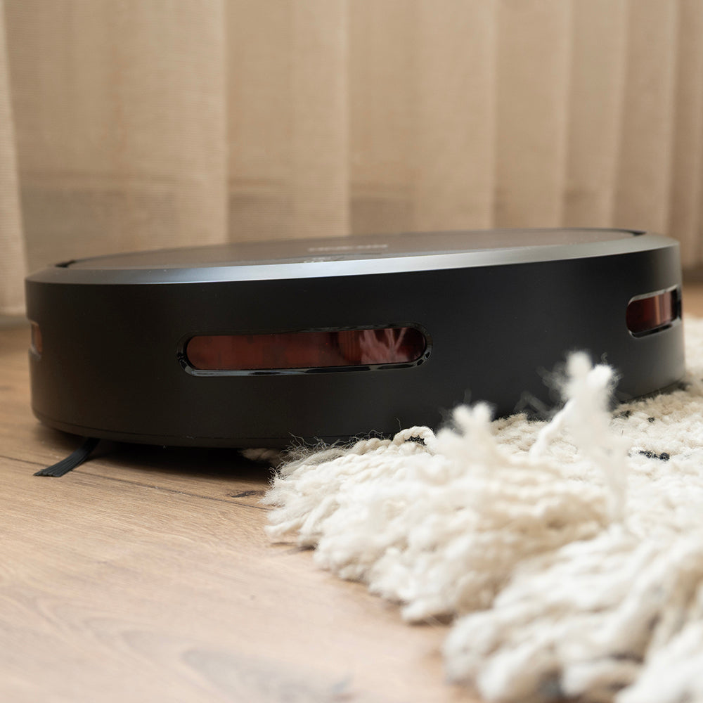 Zijaanzicht van de platte robotstofzuiger bij de franjes van een vloerkleed