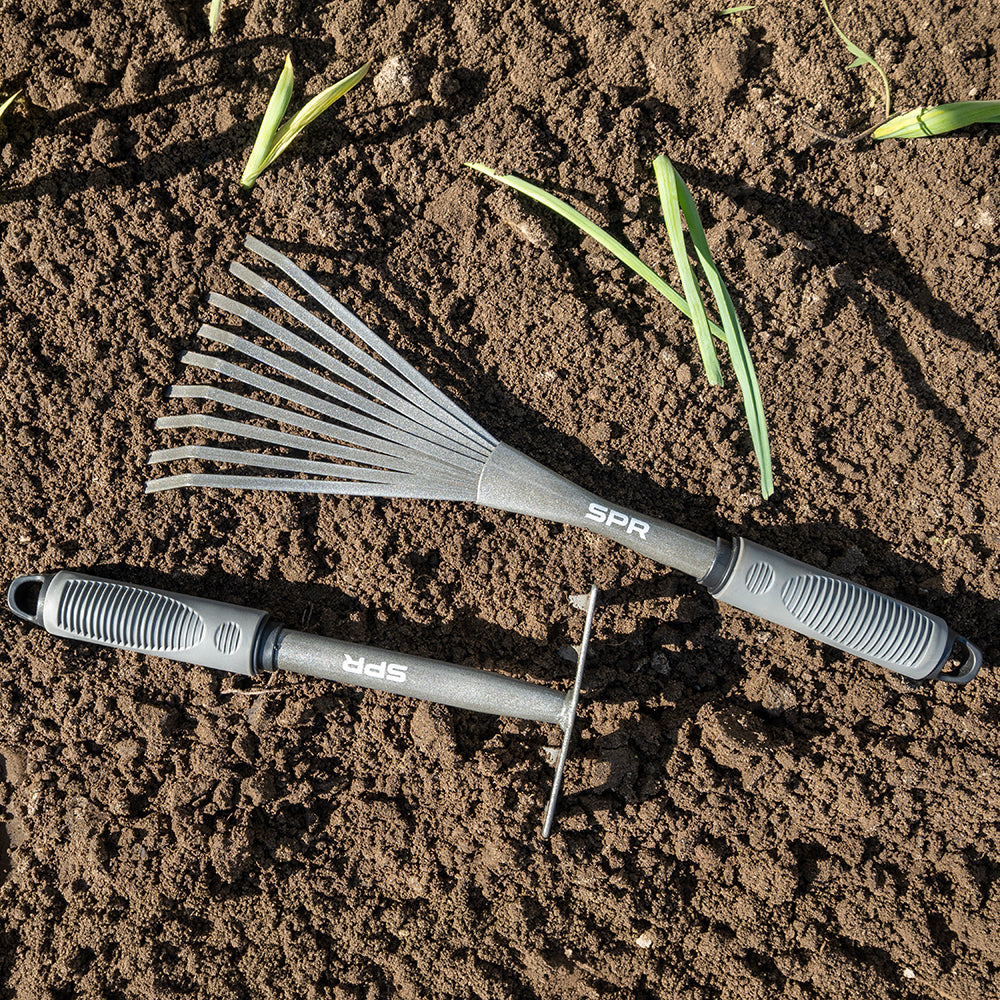 Mini-bladhark en hark met 5 tanden liggen op de aarde tussen jonge plantjes
